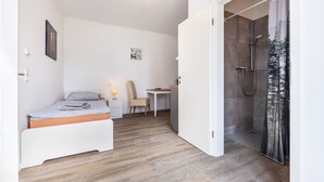 A bedroom with a bed, a desk, a chair, and a shower area.