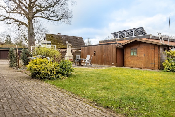 A brick pathway leading to a wooden building with a chimney, a solar panel, and a small garden with a bench.