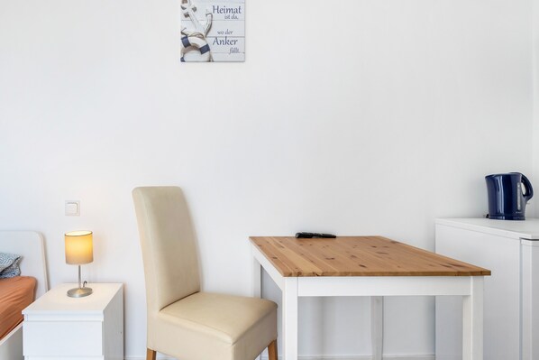 A room with a bed, bedside table with a lamp, a chair, a wooden table, and a kettle on a cabinet.