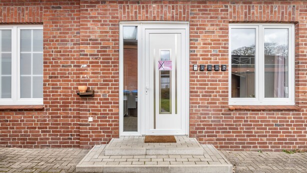 A brick building with a white door, two windows, and a small shelf with a pot.