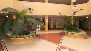 Un lobby espacioso con dos grandes palmeras en macetas circulares, un ascensor central y un piso de baldosas con un diseño geométrico.