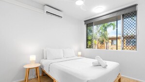 Ein minimalistisches Schlafzimmer mit einem Bett, Nachttischen, einer Klimaanlage und einem Fenster mit Blick auf Palmen.