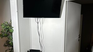 Ein Zimmer mit einem an der Wand befestigten Flachbildfernseher, einem kleinen Holztisch mit einem Korb, einem weißen Stuhl und einem Blumentopf.