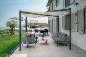 An outdoor patio area with a table and chairs, a small round table with a bottle, and a pergola providing shade. - Exclusive, elegant villa nestled among Piedmontese vineyards with a heated pool (Castiglione Tinella)
