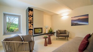 Una sala de estar con una silla de mimbre, un sofá, una mesita y vistas a un jardín a través de una ventana.