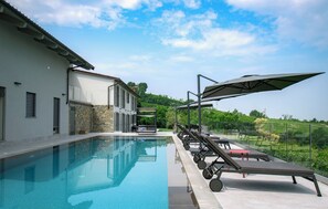 A swimming pool with lounge chairs and umbrellas, a stone wall, and a view of greenery. - Exclusive, elegant villa nestled among Piedmontese vineyards with a heated pool (Castiglione Tinella)