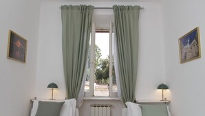 Habitació amb dos llits, una finestra amb cortines, un radiador i dues làmpades penjades a la paret.