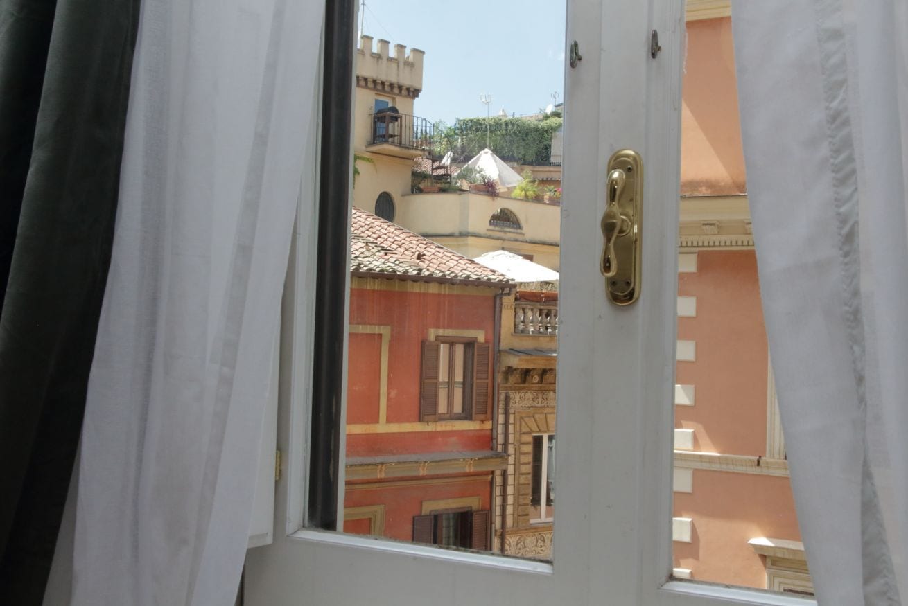 A view through an open door with a window, revealing a building with red and yellow facades, balconies, and a tiled roof.