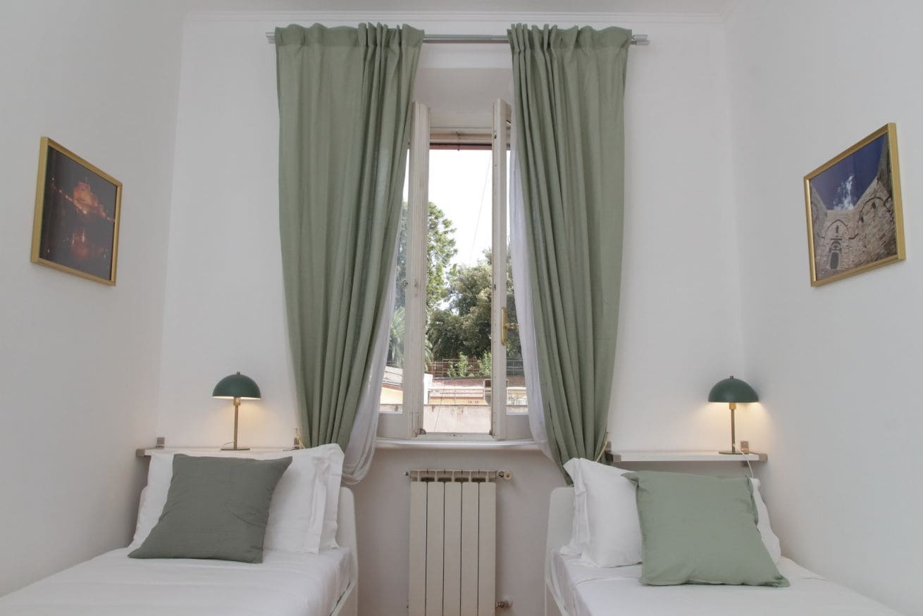 A bedroom with two beds, a window with curtains, a radiator, and two wall-mounted lamps.