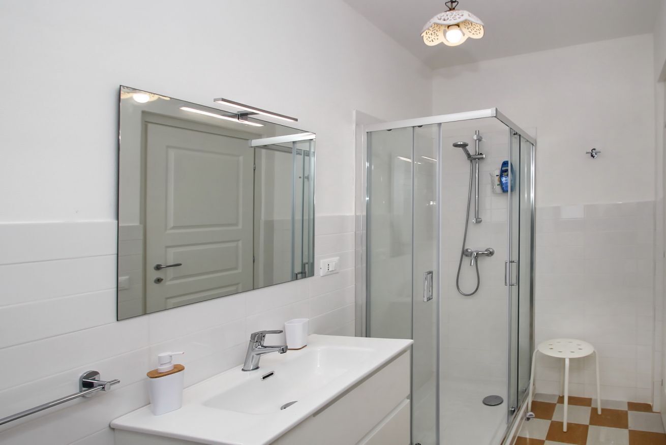 Un baño moderno con ducha de vidrio, un lavamanos blanco con un espejo encima y un taburete blanco pequeño.