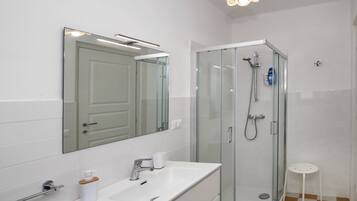 Un baño moderno con ducha de vidrio, un lavamanos blanco con un espejo encima y un taburete blanco pequeño.