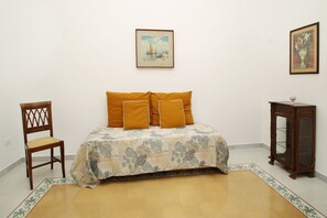 Un dormitori amb un llit, una cadira, una taula petita i dues fotografies emmarcades a la paret.