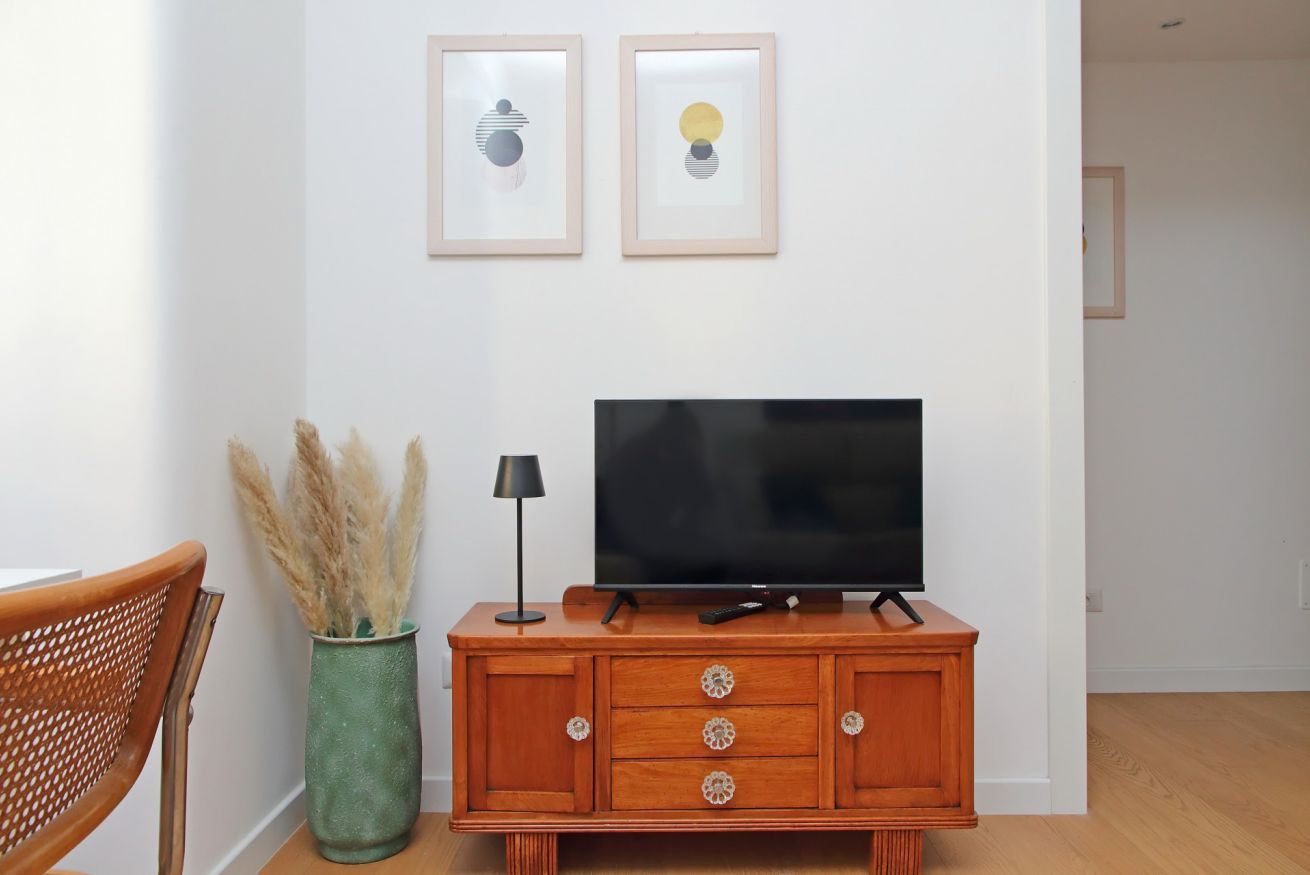 A living room with a TV on a wooden cabinet, a green vase with pampas grass, and two framed abstract artworks on the wall.