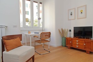 Ein Wohnzimmer mit einer Couch, einem Holzstuhl, einem kleinen Tisch, einem Fernseher auf einer Holzkommode und einem Fenster mit Blick auf Gebäude.