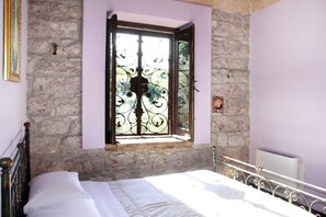 Een slaapkamer met een stenen muur, een bed met wit beddengoed, een raam met decoratieve ijzeren spijlen en een klein schilderij aan de muur.
