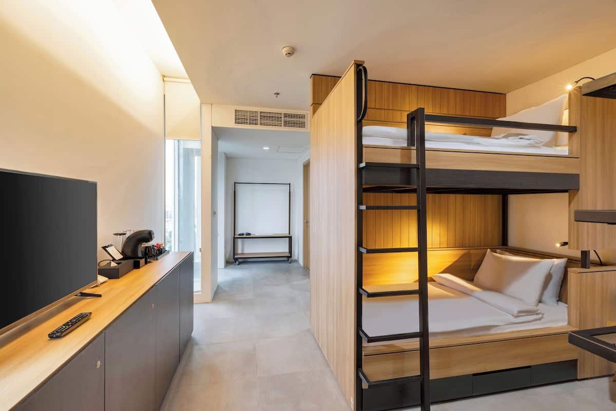 A modern hotel room with a bunk bed, a desk with a flat-screen TV, and a small seating area.