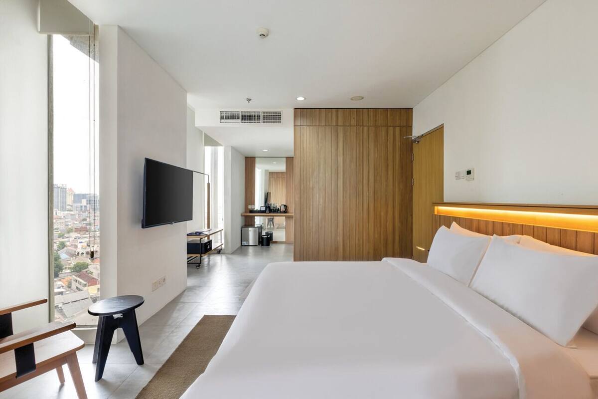 A modern hotel room with a large bed, a flat-screen TV mounted on the wall, a small stool, and a view of the city through a large window.