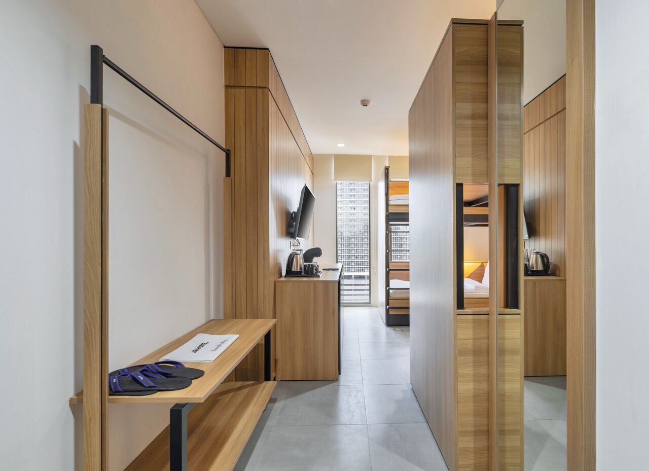 A modern hotel corridor with wooden paneling, a bench with a paper, a flat-screen TV, and a kitchenette area.