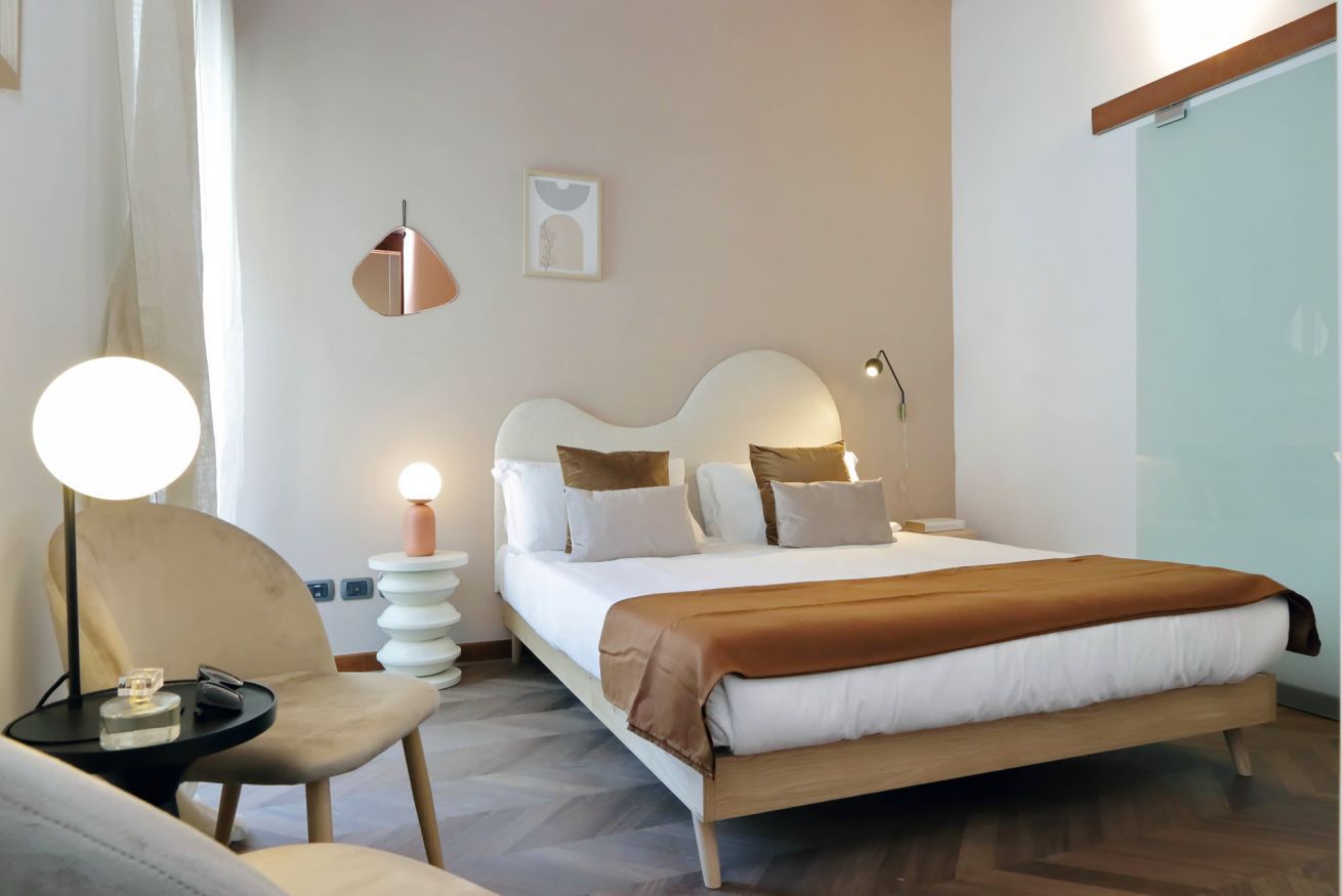 A modern hotel room with a large bed, a bedside table with a lamp, a chair, and a wall-mounted picture.