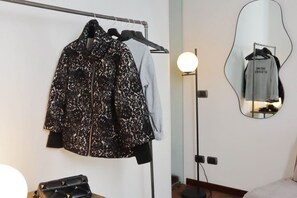Een kamer met een kledingrek waarop een zwarte jas met bloemenprint en een grijs shirt liggen, een ronde spiegel die de kamer weerspiegelt, en een zwarte handtas op een tafel.