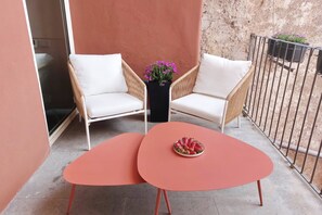 Een balkon met twee rieten stoelen, een tafeltje met een schaal fruit en een bloempot met paarse bloemen.