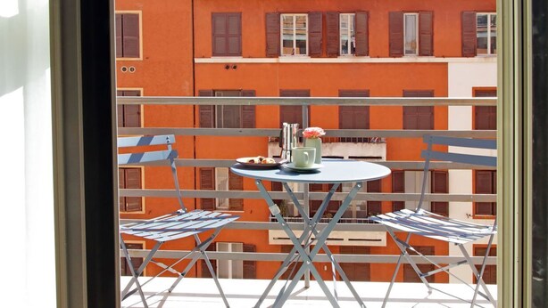 En balkon med et bord og stole, en vase med blomster, og en bygning med orange vægge og skodder i baggrunden.