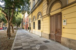 A long, straight sidewalk with a checkered pattern, flanked by trees and buildings with large wooden doors and windows. - Cami’s Corner - Heart of Cagliari (Cagliari)
