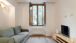 A living room with a green sofa, a TV on a wooden stand, a window with a view of trees, and a white radiator. - Cami’s Corner - Heart of Cagliari (Cagliari)