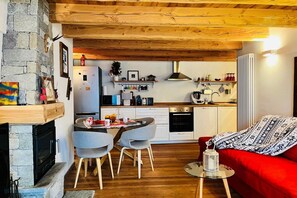 A cozy interior with a stone fireplace, wooden ceiling beams, a dining area with chairs, a red sofa, and a kitchen with white cabinets and appliances.