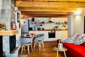 A cozy interior with a stone fireplace, wooden ceiling beams, a dining area with chairs, a red sofa, and a kitchen with white cabinets and appliances.