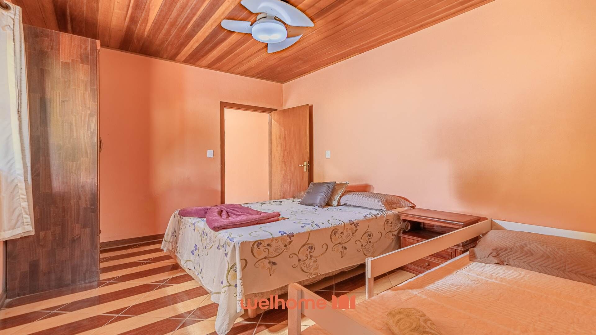 A bedroom with a wooden ceiling, a bed with a floral bedspread, a wooden wardrobe, and a door.