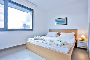 A modern hotel room with a large window, a bed with neatly folded towels, a bedside table with a lamp, and a framed picture on the wall.