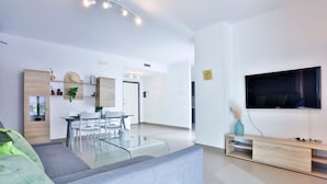 Una sala moderna con un sofá gris, una mesa de centro de vidrio, una zona de comedor con una mesa y sillas blancas, un televisor montado en la pared y un mueble de televisión de madera.