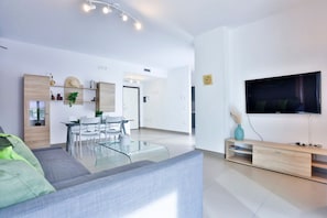 A modern living room with a grey sofa, a glass coffee table, a dining area with a white table and chairs, a TV mounted on the wall, and a wooden TV stand.