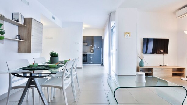 Una sala moderna con una mesa de comedor de vidrio, sillas blancas, un televisor de pantalla plana y una cocina al fondo.
