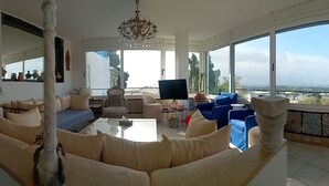 A spacious living room with a large window, a chandelier, and a view of the city. - Maison L'étoile de Sidi Bou Saïd - 360° Sea View (Sidi Bou Said)