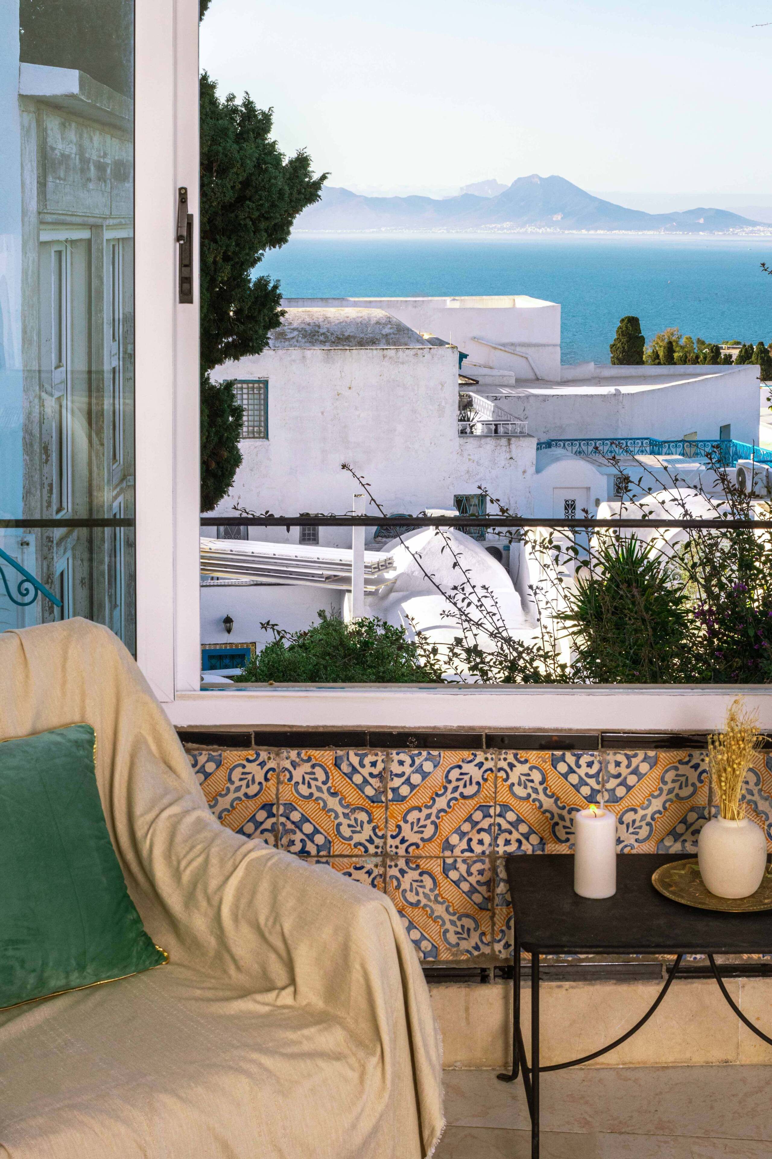 A balcony with a view of the sea and white buildings, a beige sofa, and a patterned tile backsplash.