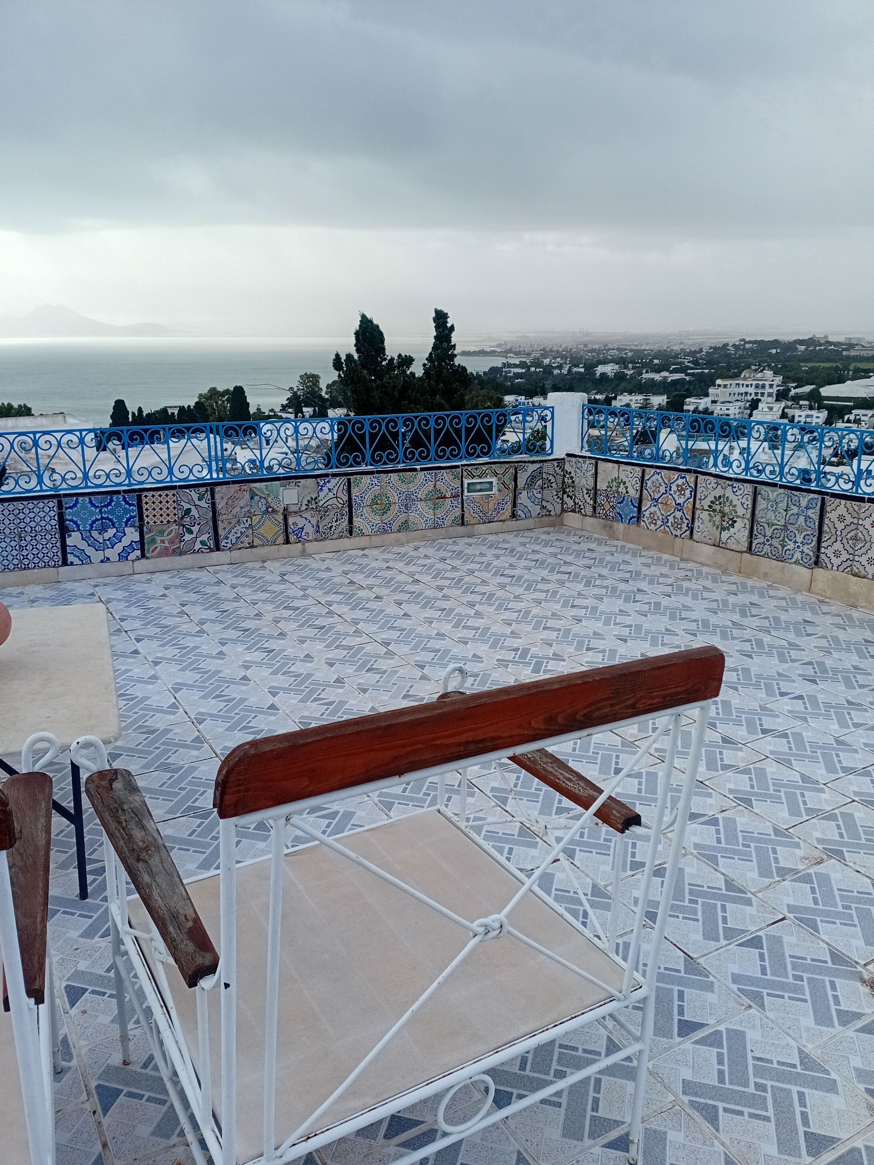 Une terrasse avec des carreaux décoratifs, un banc en bois et une vue sur la mer et les bâtiments.