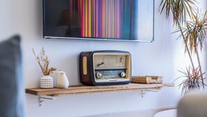 Une vieille radio posée sur une étagère en bois, un vase contenant des plantes séchées et une œuvre d’art colorée à rayures accrochée au mur.