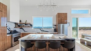 A modern kitchen with a marble backsplash, wooden cabinets, a central island with bar stools, and large windows offering a view of the desert landscape. - Desert Seclusion offers Pickleball, Pool, Spa, incredible stargazing (Joshua Tree)