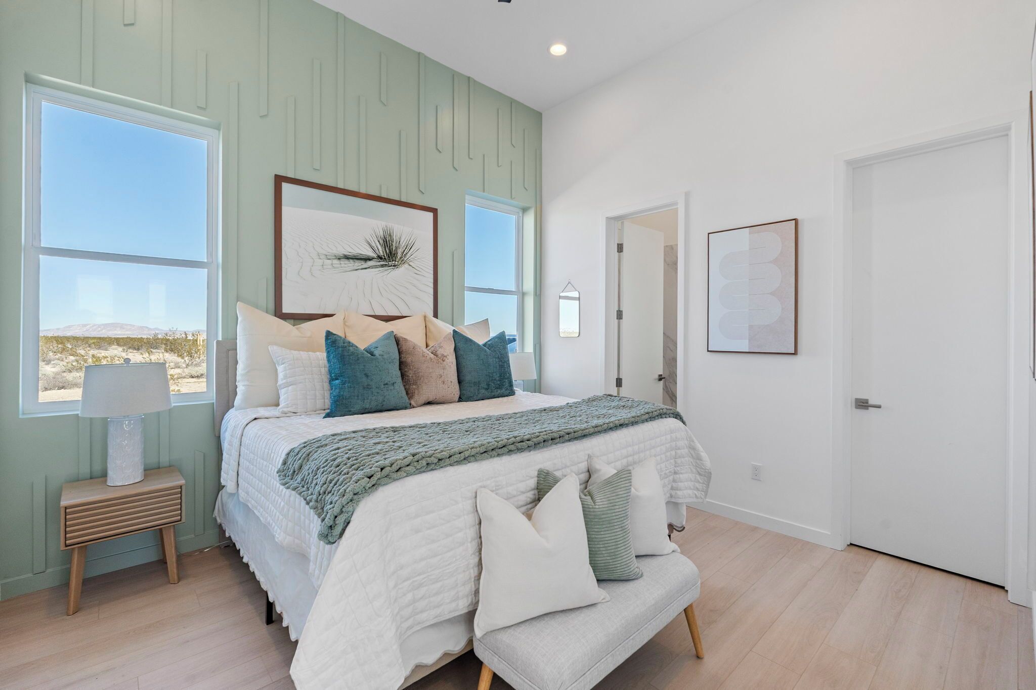 A bedroom with a large bed, a nightstand, a lamp, a framed artwork, and a view of the desert through the window.