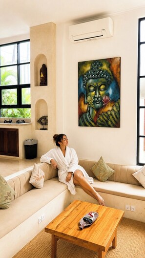 A person in a white bathrobe sitting on a beige sofa in a room with a large Buddha painting, a wooden coffee table, and a window with a view of greenery. - 3BR Private Pool Villa in Berawa • Walk to Beach, Cafes & Finns (Bali)