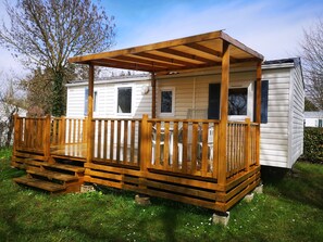 Une terrasse en bois avec une pergola, un petit porche et une maison mobile blanche avec des volets.