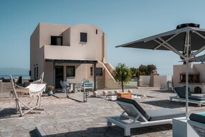 A modern building with a flat roof and a patio area with lounge chairs and a hammock. - Mythique Pool Residence - Oia Santorini (Oia)