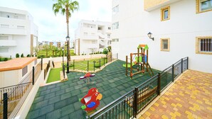 A children's play area with a slide, swings, and a climbing structure, surrounded by a white building with balconies and a palm tree. - Linnea Sol by Villas&You (Orihuela Costa)