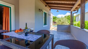 Eine überdachte Terrasse im Freien mit Korbmöbeln, einem Tisch mit Obstschale und Weingläsern sowie Blick auf Grünflächen und einen Pool im Hintergrund.