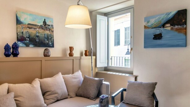 Una sala de estar con un sofá, una mesa redonda y dos sillas. Hay cuadros en las paredes, una ventana con vistas y una lámpara.