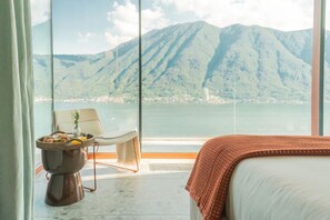 A hotel room with a large window, a bed, a chair, and a small table with a basket of bread. - Villa Aurea - My Home in Como (Sala Comacina)