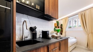 Een compacte keuken met een zwarte spoelbak, houten kastjes en een marmeren achterwand. Er is een koffieapparaat, een plant en een bed zichtbaar in de aangrenzende kamer.