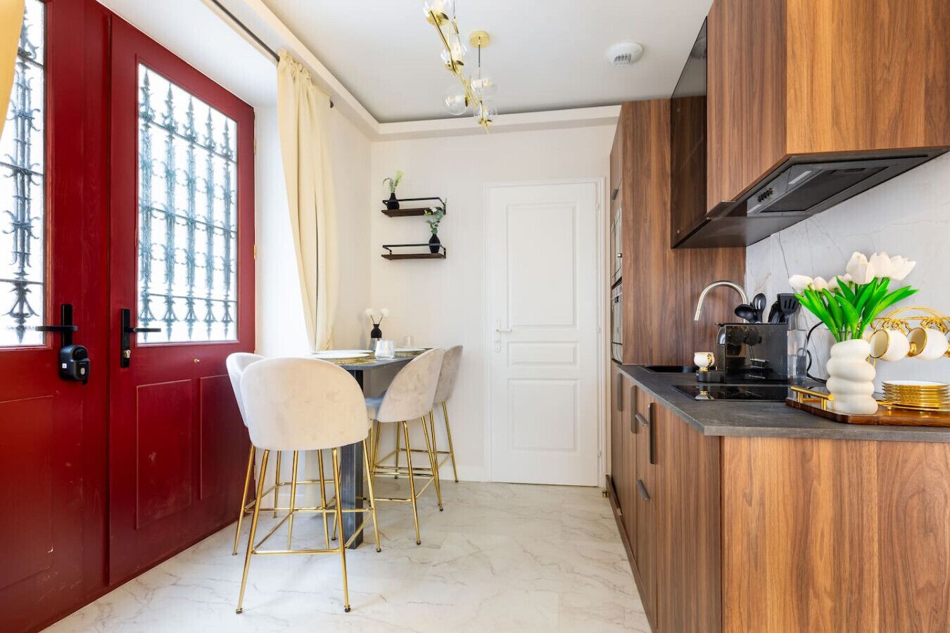 Una cocina moderna con una puerta roja, paredes blancas y un suelo de mármol. Hay un rincón comedor con dos sillas blancas y una mesa.